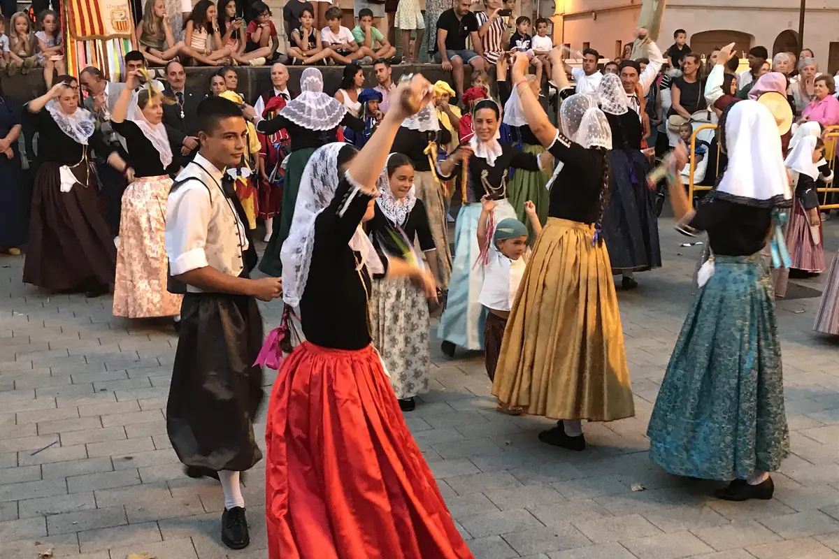 Bailarines con traje tradicional, mujeres con pañuelo de encaje blanco en la cabeza y falda larga roja, bailando en una plaza empedrada con espectadores detrás