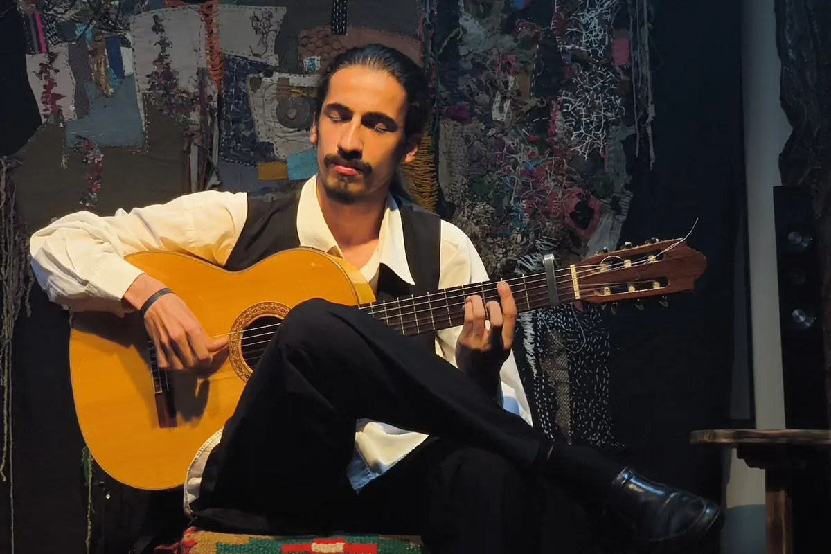Ein sitzender Gitarrist im Westen, der vor einem gemusterten Stoffhintergrund eine spanische Gitarre spielt